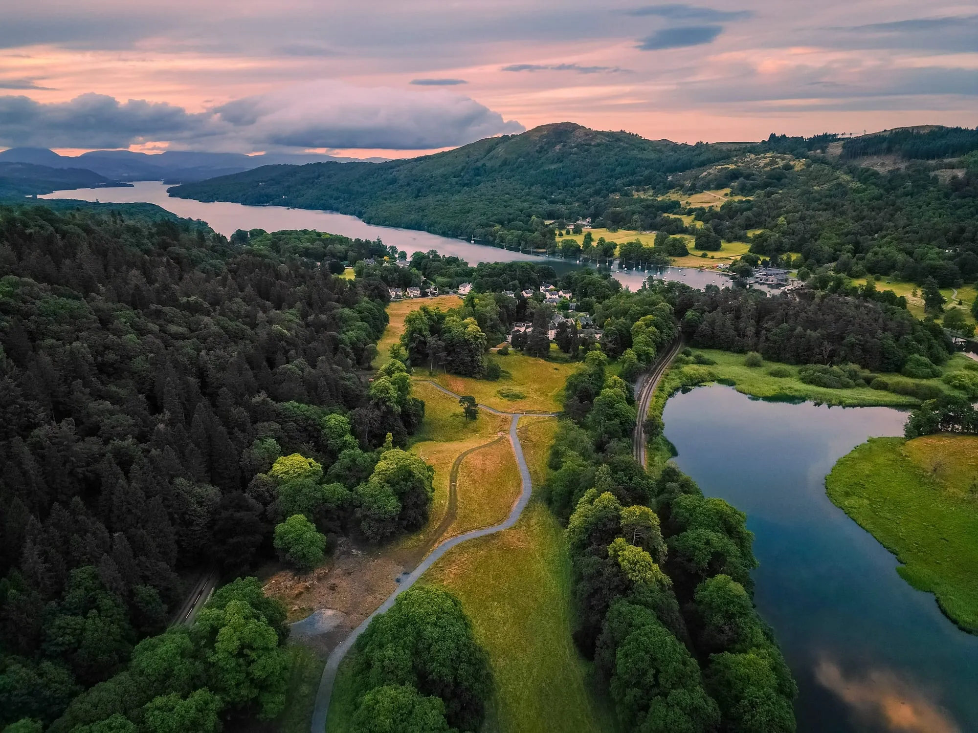 Aerial view at dusk of a lake winding between wooded hills, a path curving through parkland under a pink and grey sunset sky