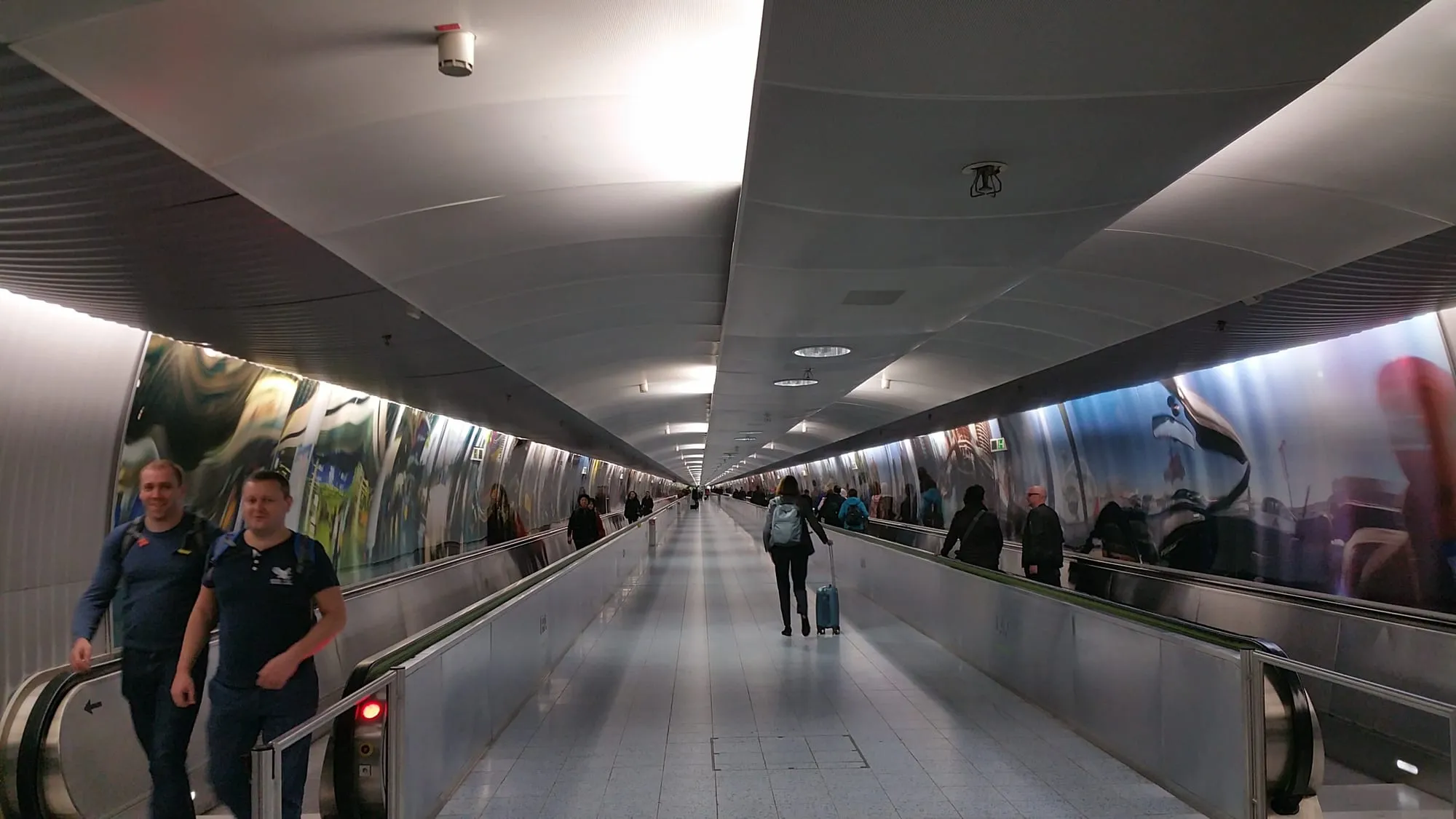 A long airport pedestrian tunnel lined with moving walkways and mirrored panels, travellers passing beneath a curved white ceiling