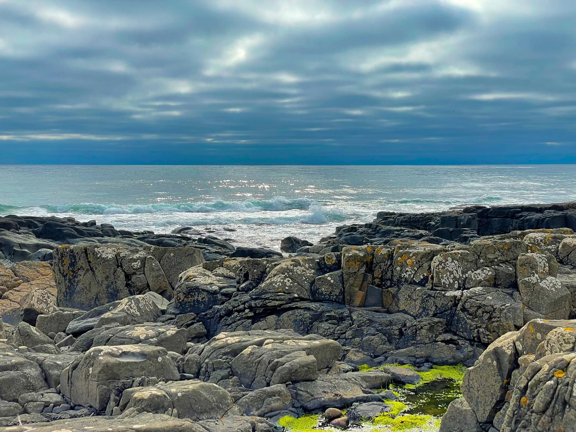 Waves breaking beyond a rugged, lichen-covered rocky shoreline under a heavy grey-blue sky, with green weed in a foreground pool