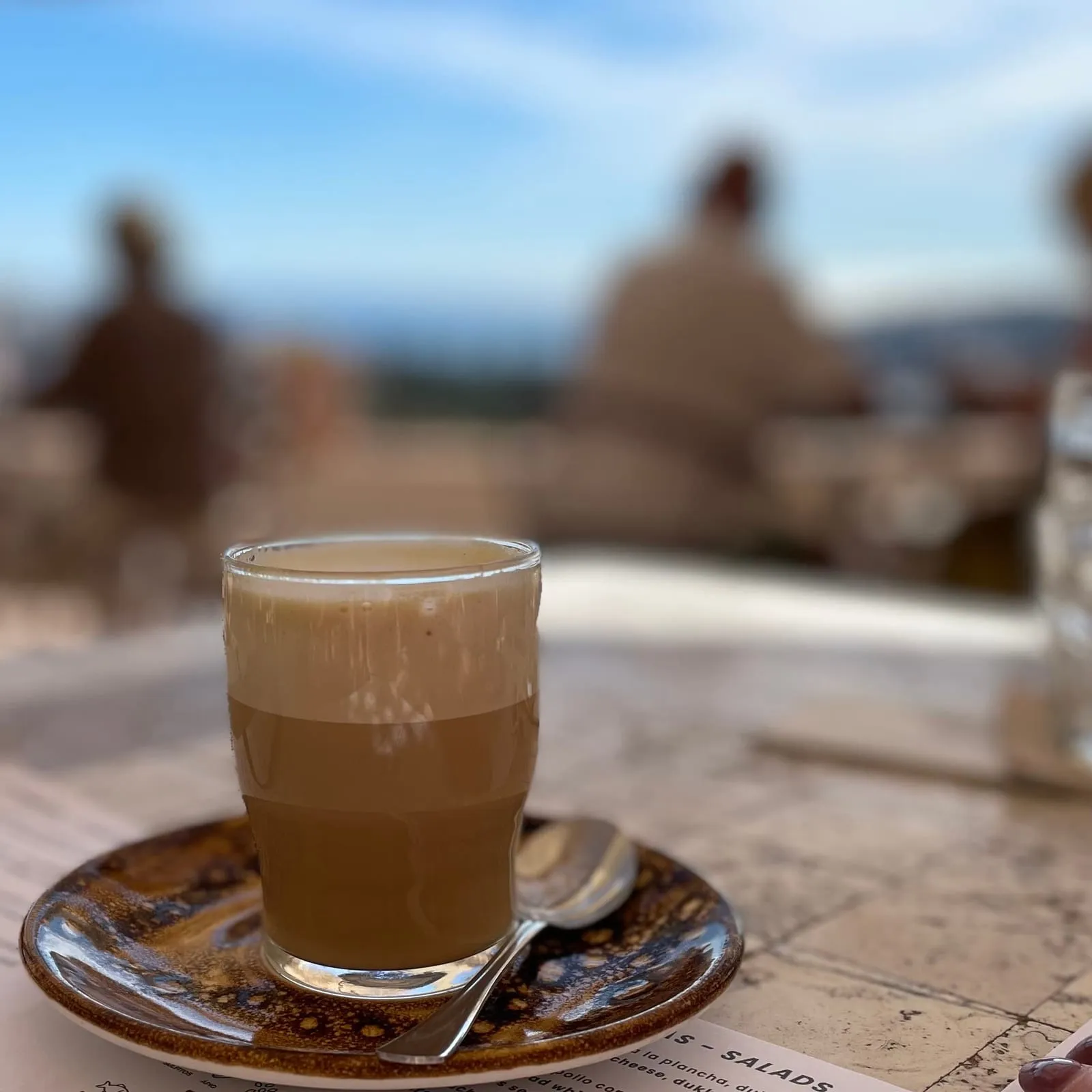 A glass of milky coffee on a glazed brown saucer with a teaspoon, on a sunlit terrace table with a hazy coastal view behind