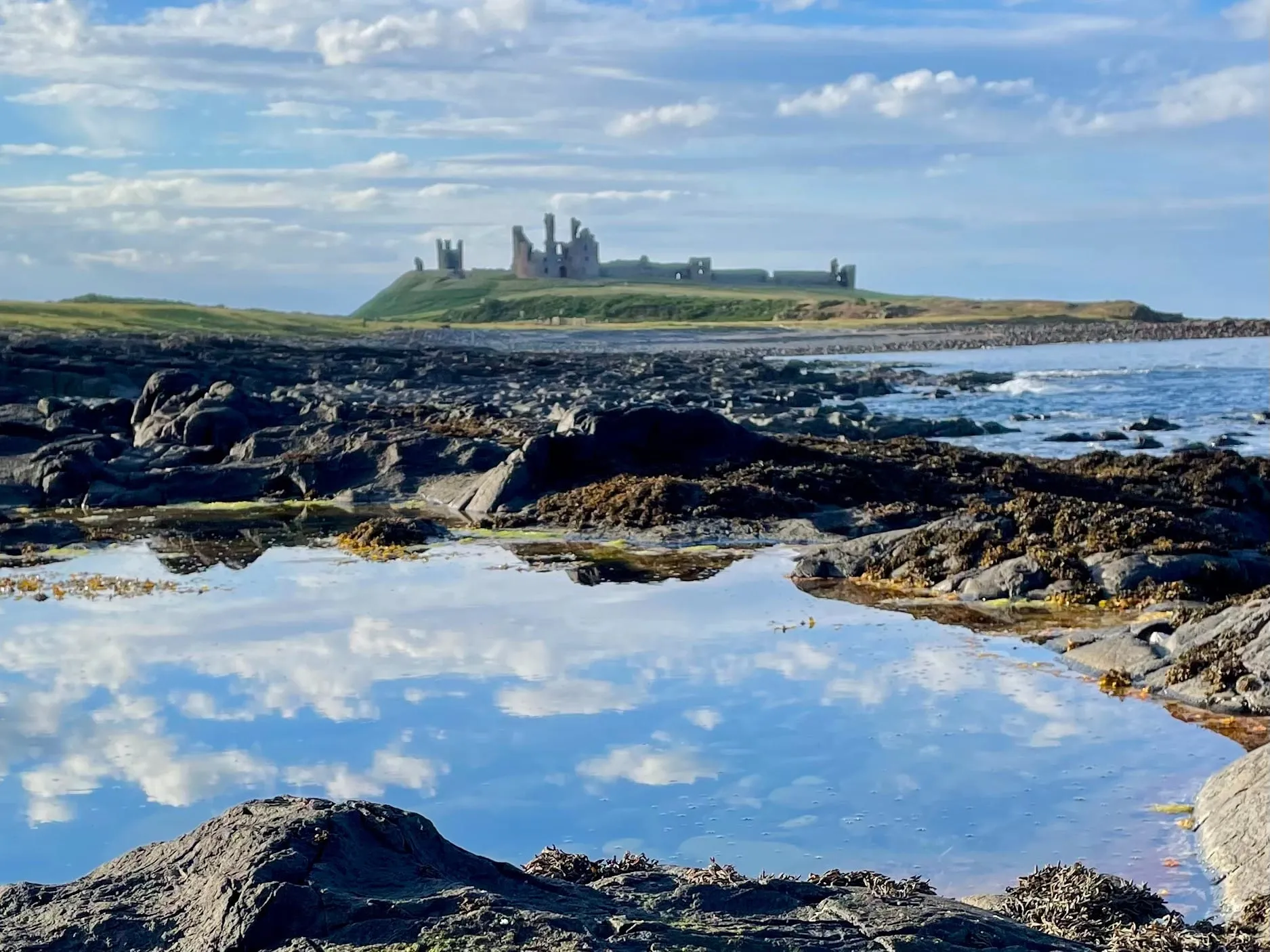 The ruins of Dunstanburgh Castle on a low headland, seen across a rocky shore with a still tidal pool reflecting the sky