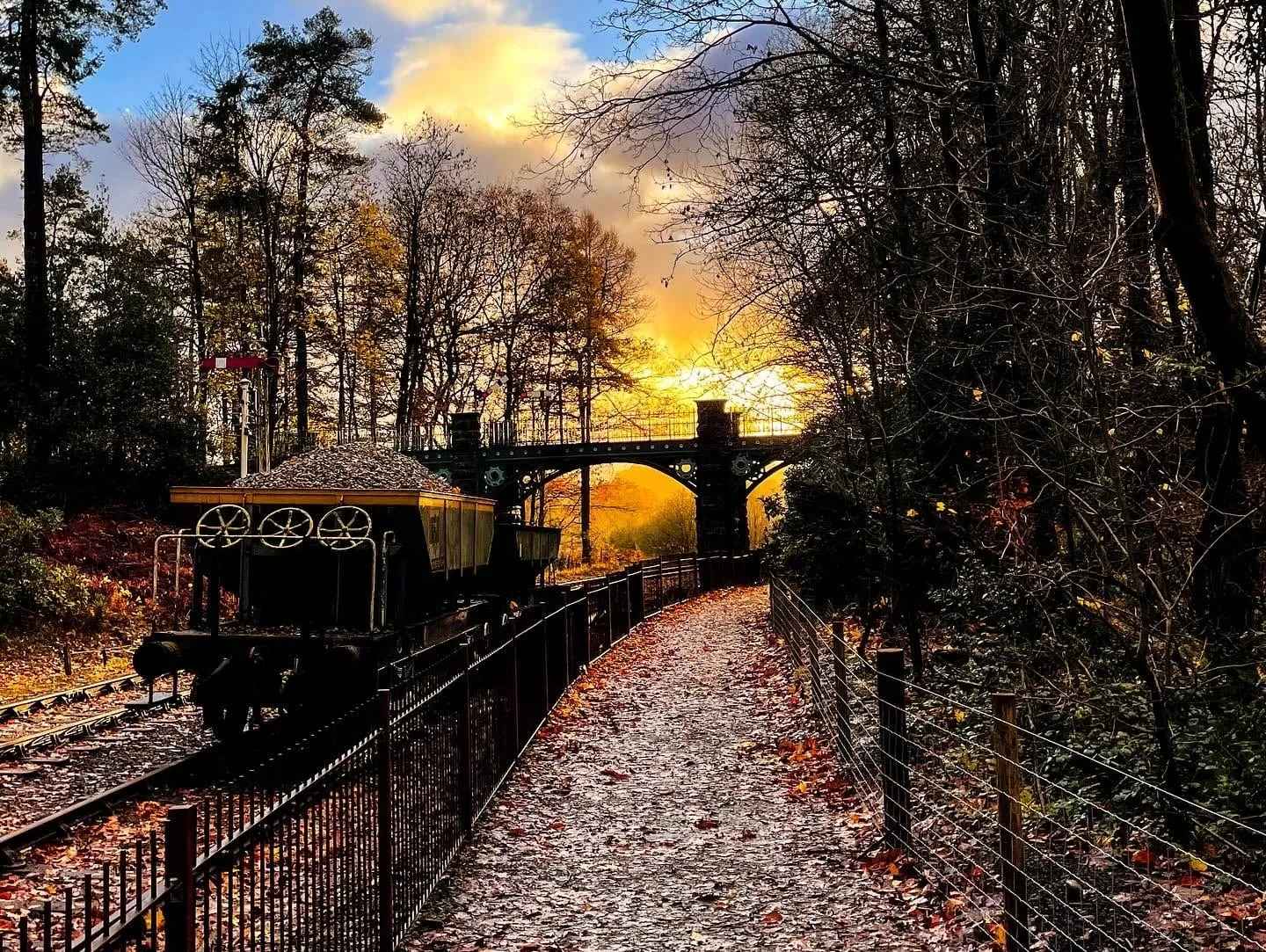A miniature railway wagon beside a leaf-strewn path, with an arched viaduct and a golden autumn sunset behind bare trees