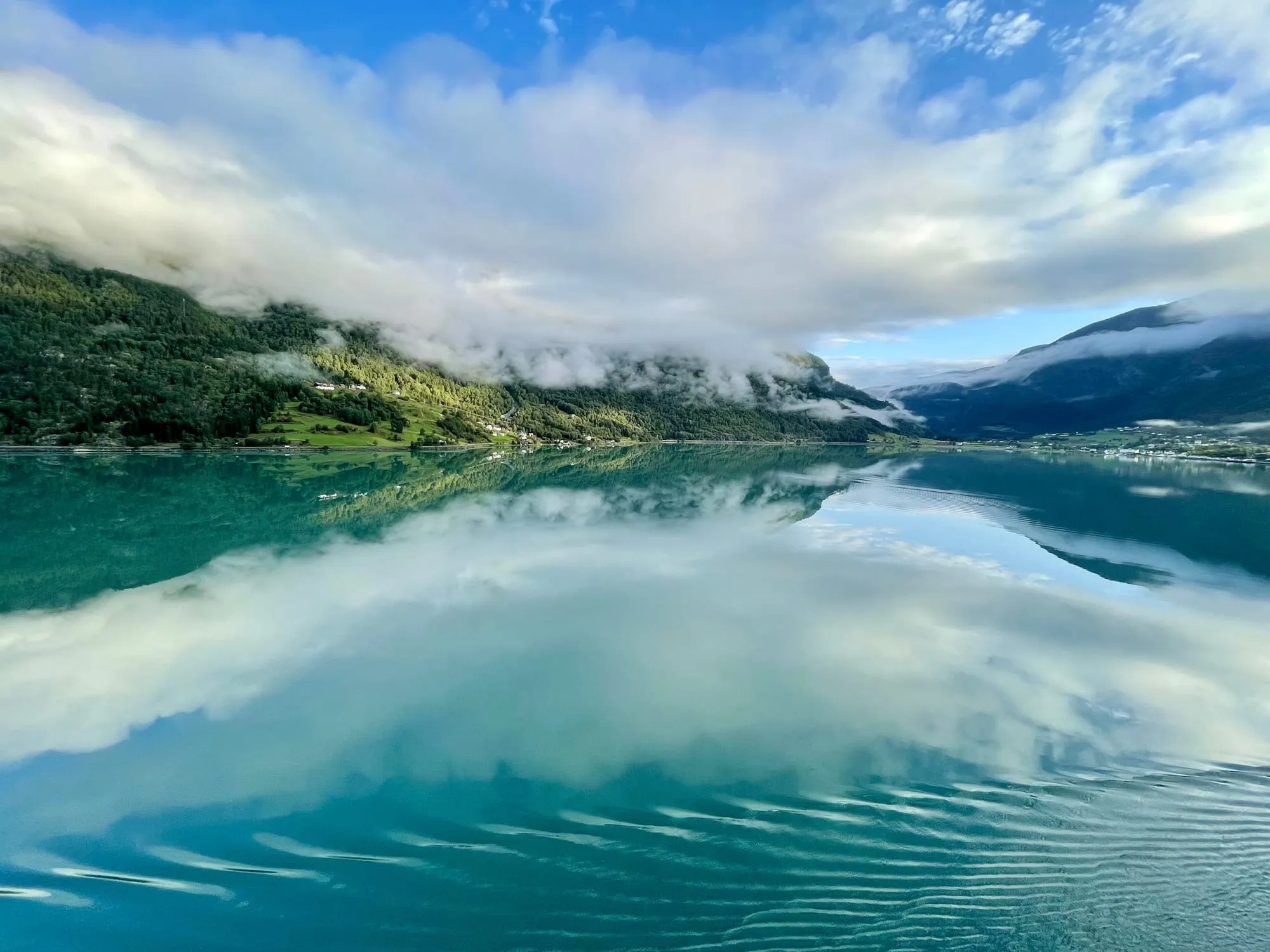 A calm Norwegian fjord, turquoise water mirroring clouds and forested mountainsides under low, drifting cloud