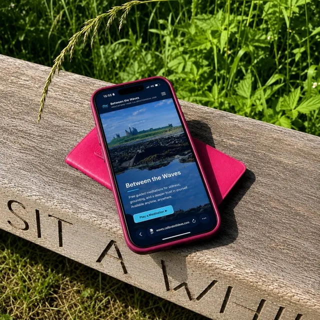 The Between the Waves app open on a phone resting on a weathered wooden bench carved with the words 'Sit a while', among sunlit summer grass