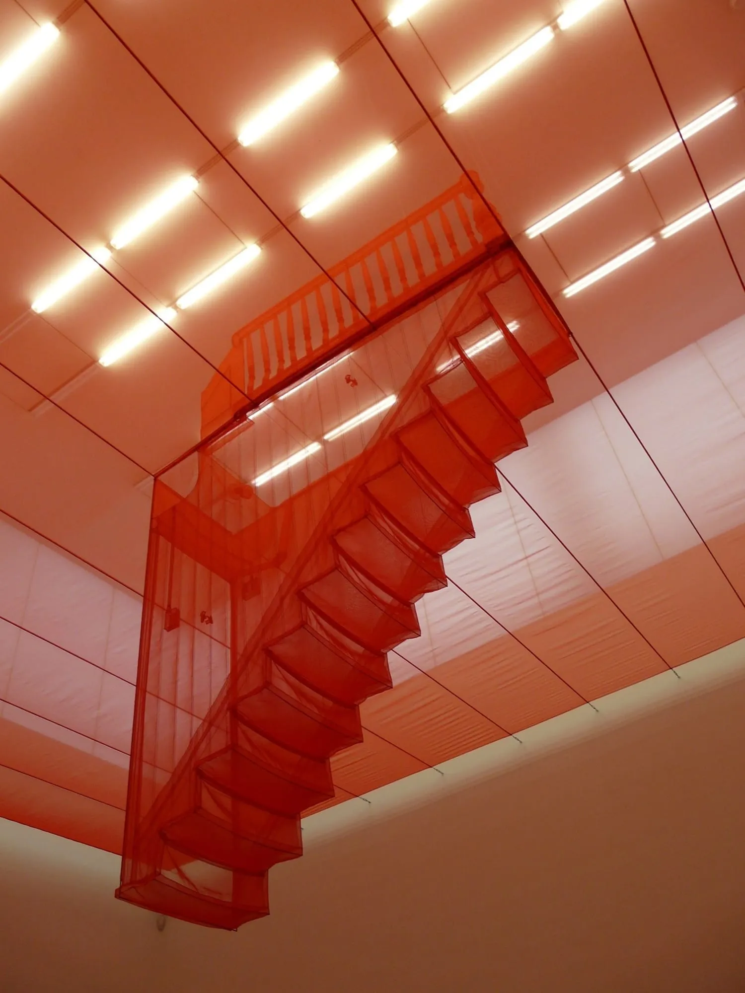 A staircase sculpted in translucent red fabric, suspended beneath a gallery ceiling and lit from above